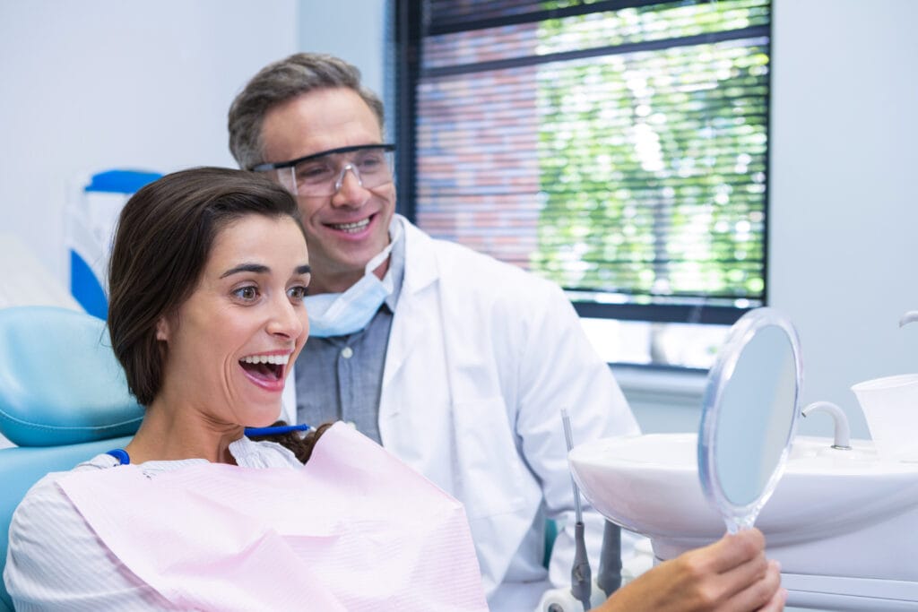happy woman looking at mirror by dentist while sitting on chair at medical clinic