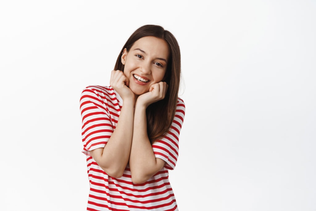portrait of cute brunette woman smiling, tilt head and gazing with admiration, looking adorable, holding hands near face and grinning happy, standing against white background